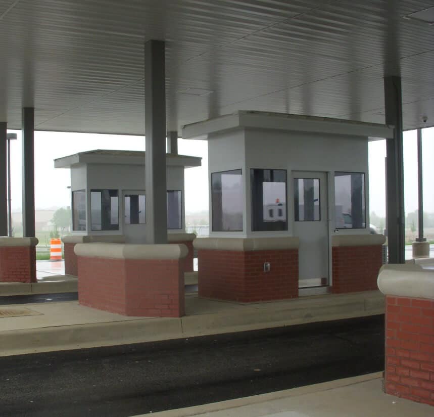 Two small, brick toll booths with grey roofs under a large metal canopy, beside a wet, empty toll road on a cloudy day. The booths are Porta-King prefabricated structures designed to