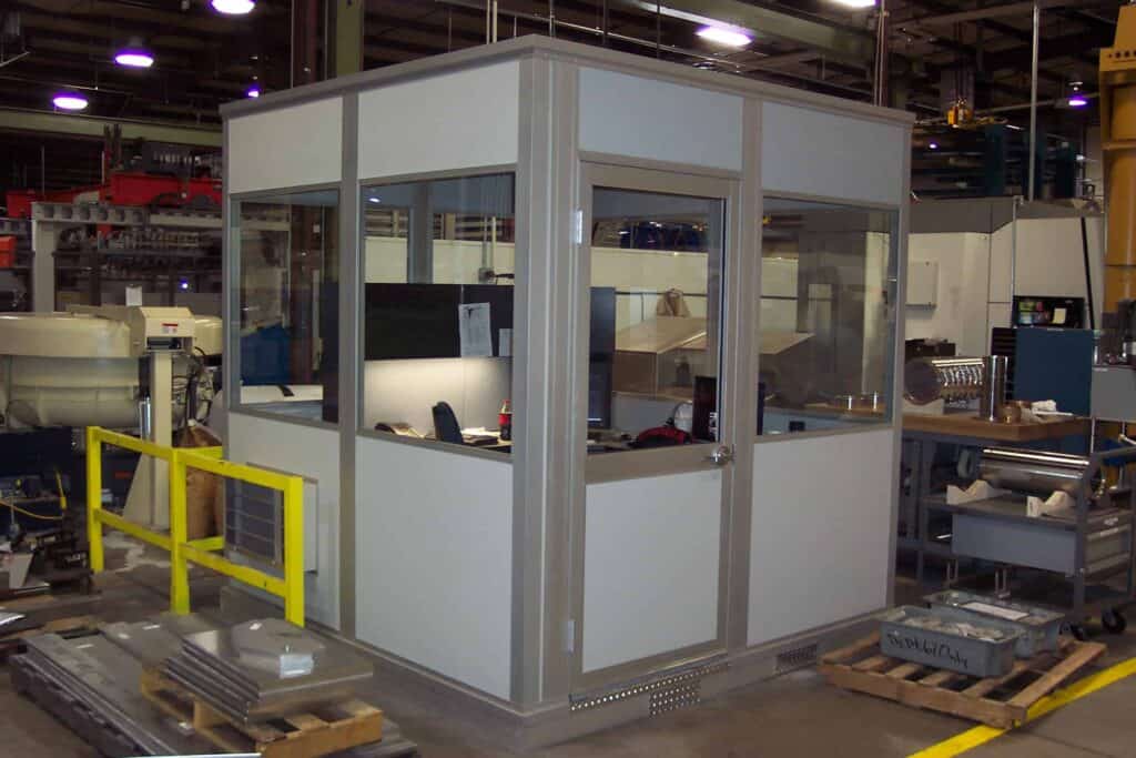 A small, modern office cubicle with glass walls situated in an industrial factory setting, surrounded by machinery and equipped with modular cool rooms to safeguard employees.