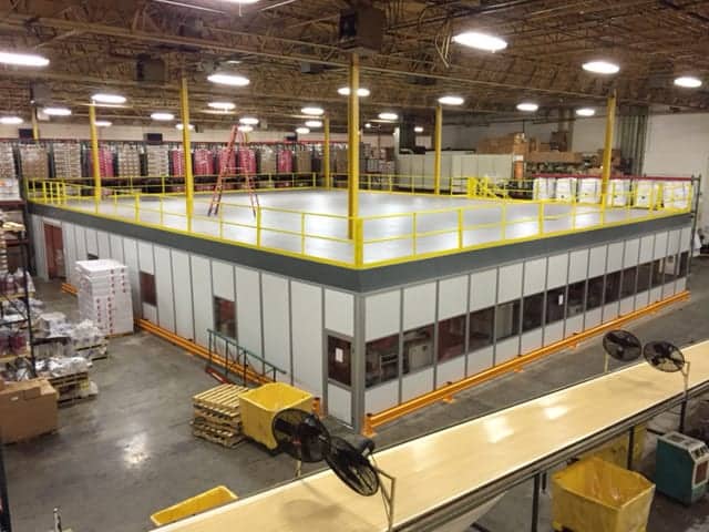 A well-lit warehouse interior showing a two-level inplant office space with glass windows and yellow safety railings, surrounded by industrial shelves stocked with boxes and supplies.