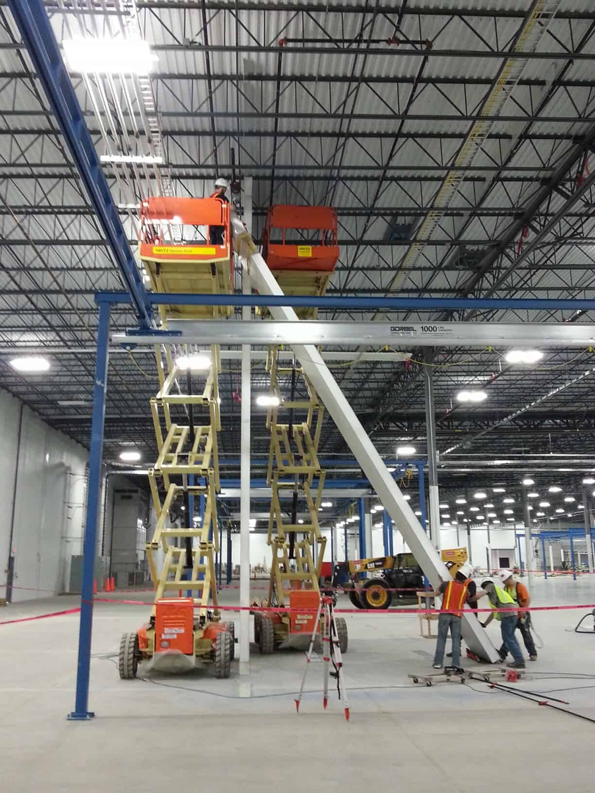 Two workers in a warehouse using scissor lifts to access high areas. One lift is extended towards the ceiling. They wear safety gear, and another group discusses plans below near modular partition walls.