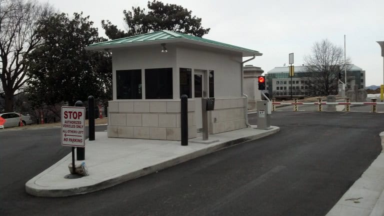 A security checkpoint booth with a stop sign and red traffic light at a gated entrance, featuring a freshly paved road and overcast sky. This setup includes essential additions typical of prefabricated military guard sh