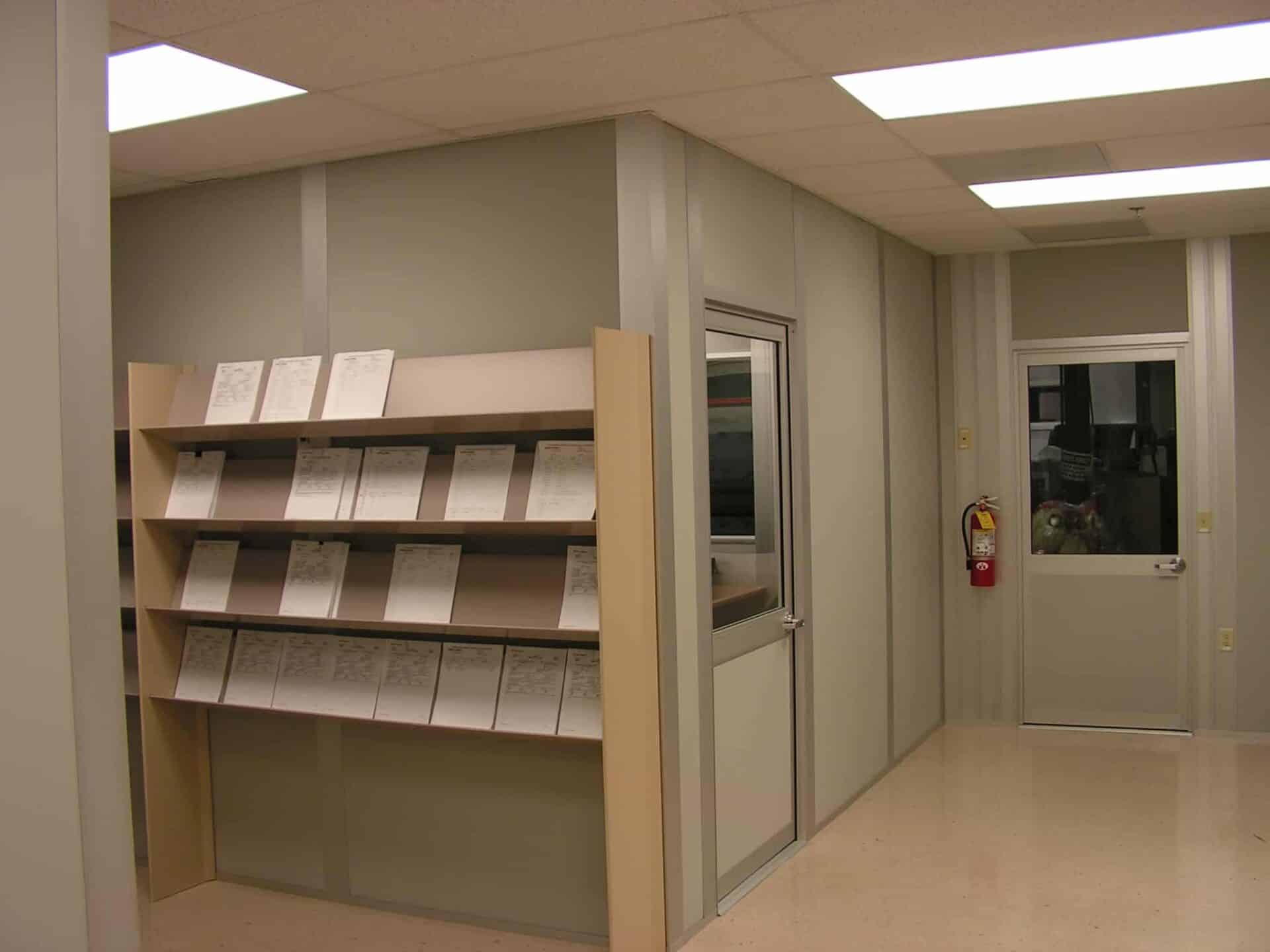 A clean, well-lit office corner in the new wing of the hospital with a large shelf filled with neatly organized documents. There's a glass door on the right and a fire extinguisher mounted on