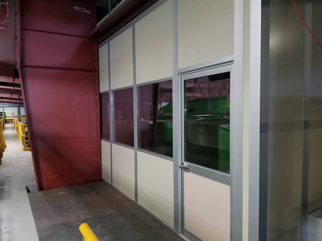 An industrial setting with a metallic structure and tinted glass modular rooms, showcasing internal green lighting. A maroon wall is visible on the left, with exposed conduits and a yellow barrier in the background