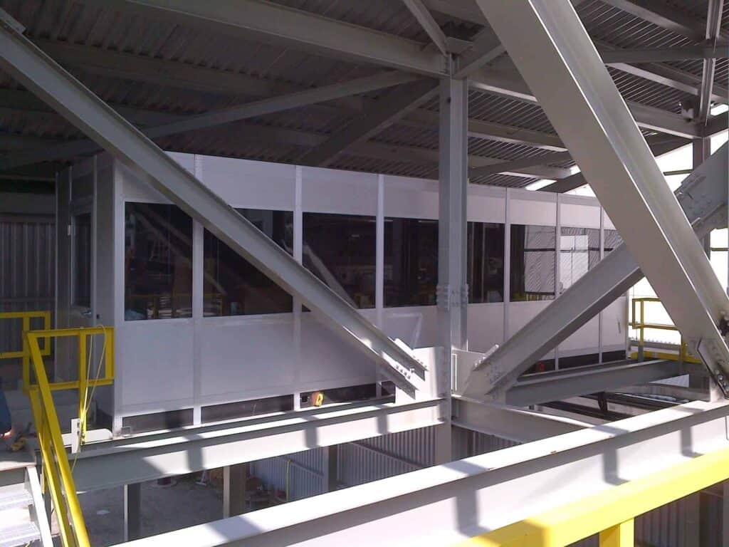 Interior of an industrial facility with large metal beams and structural supports visible. The space includes gray walls, bright yellow safety barriers, operator booths, and a clear view of construction elements.