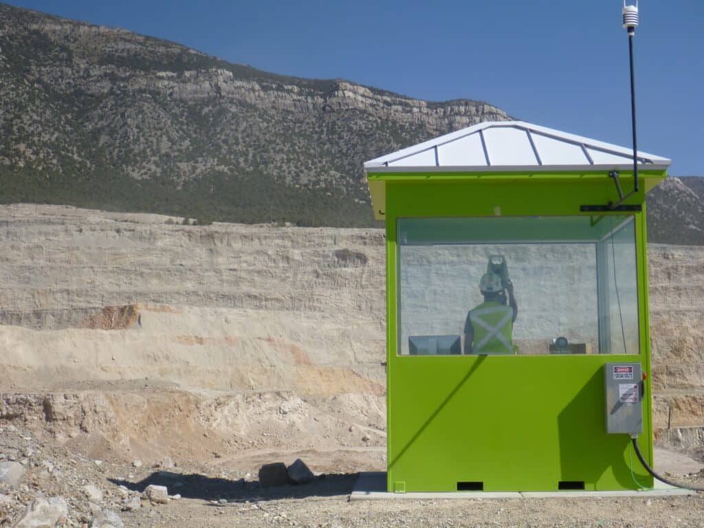 A bright green prefabricated security booth with large windows and a white roof, located in front of a large, layered rock quarry under a clear blue sky.