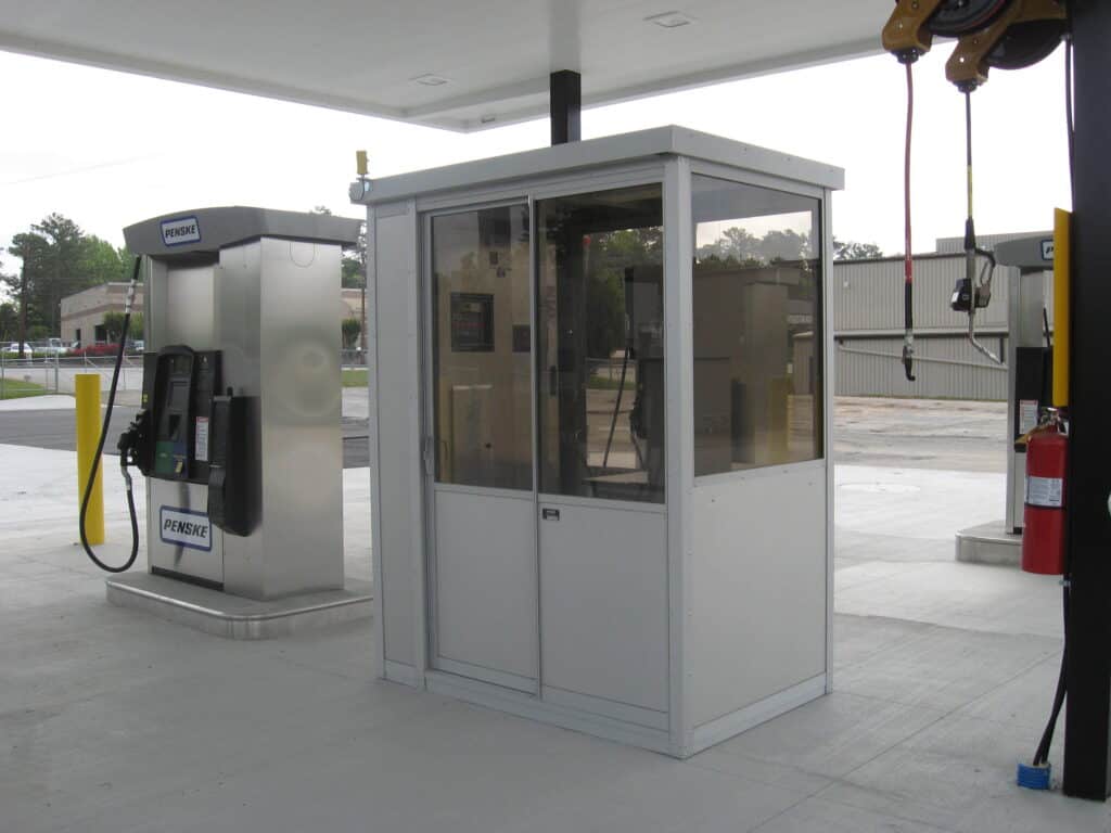 A small prefabricated control room made of glass and metal, located under a shelter at a parking facility, equipped with a payment terminal and intercom system.