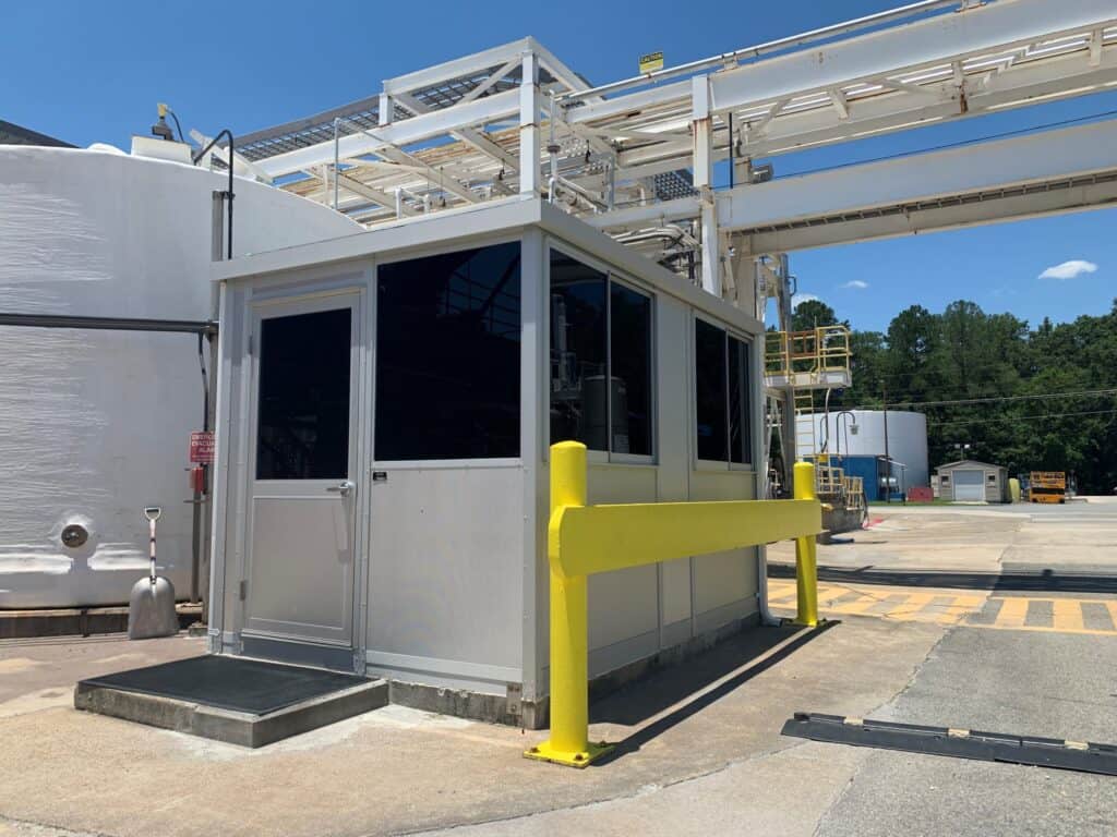 A small industrial building with large windows and a yellow guardrail, situated under a metal framework, housing prefabricated control rooms, with tanks and equipment in the background under a clear sky.