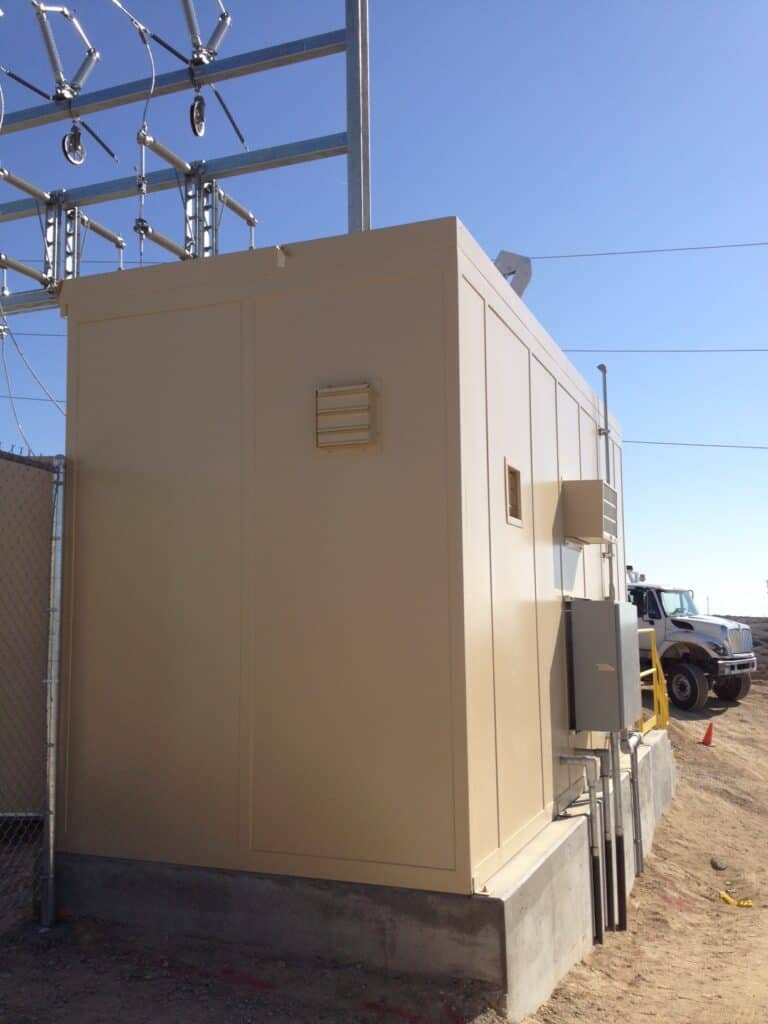 Switch Gear Enclosure 1 A large beige electrical switchgear enclosure with ventilation grilles, situated in a fenced power station area under a clear sky. A truck can be seen in the background.