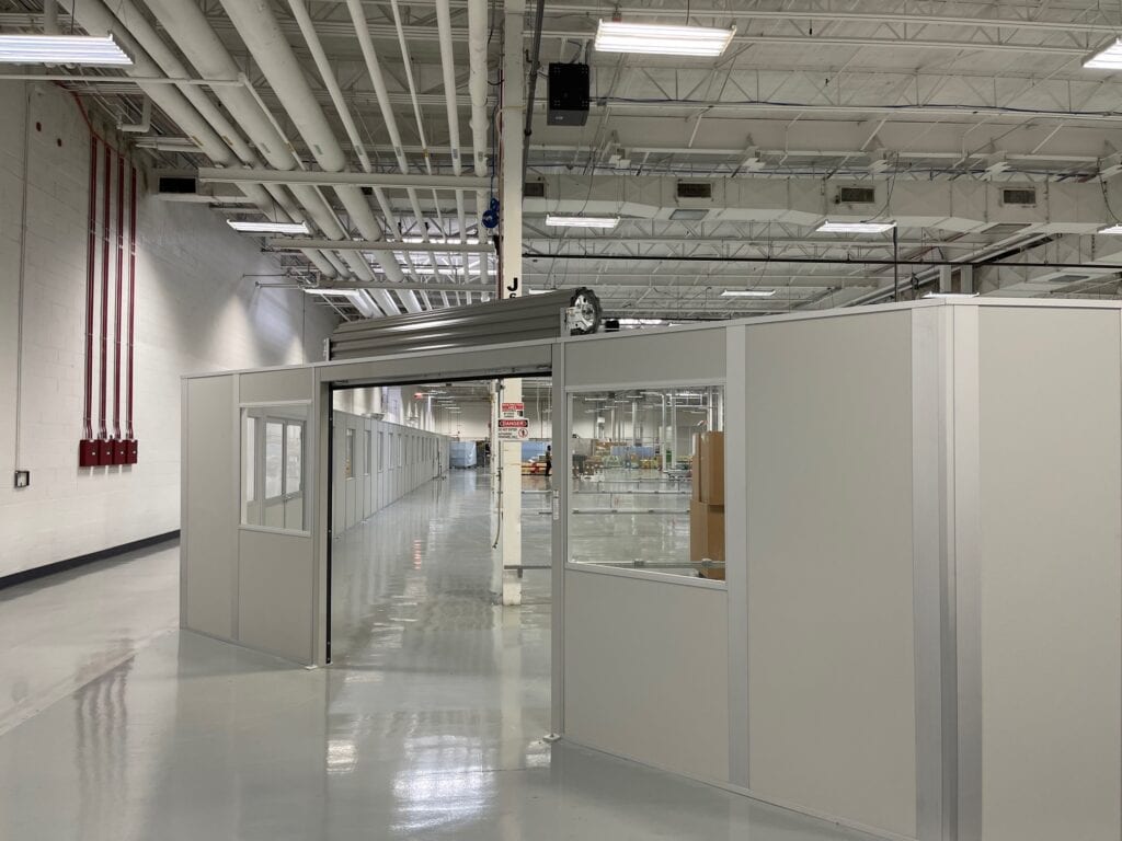 Interior of a modern industrial facility featuring glossy floors, high ceiling with exposed pipes, and glass-walled cubicles along a long corridor with freestanding partitions.