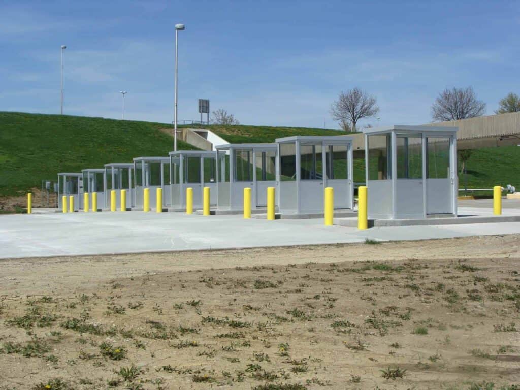 A row of new, modern bus shelters with transparent walls and metal roofs, lined up on a freshly paved area with yellow bollards, on a sunny day near toll booths.