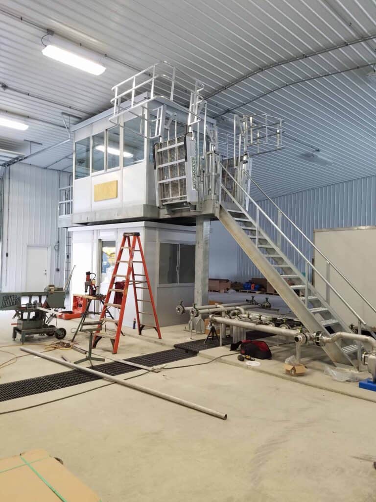 An industrial facility with a metal staircase leading to a raised control room with operator booths. Tools and construction materials are scattered around the spacious, well-lit interior.