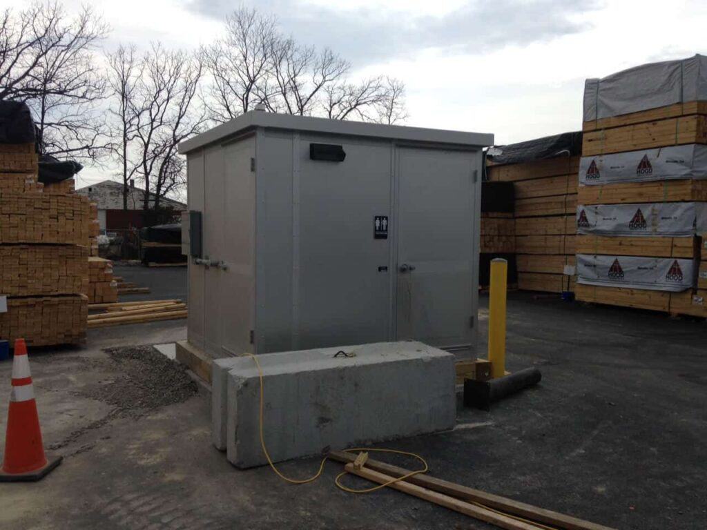 A small, gray utility shed located in a commercial area with stacks of wooden pallets and boards in the background. There are yellow bollards near the shed and portable outdoor restrooms nearby under a