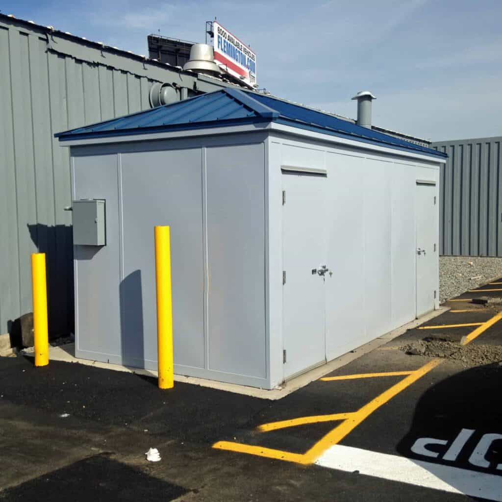 A gray industrial outdoor electrical utility building enclosed by a chain-link fence, with safety yellow bollards and portable restrooms in front, under a clear blue sky above.