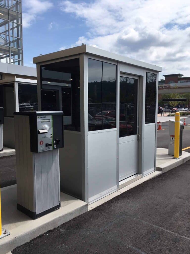 A parking lot attendant booth with glass windows and an adjacent ticket dispenser at the entrance of a sunny parking lot.