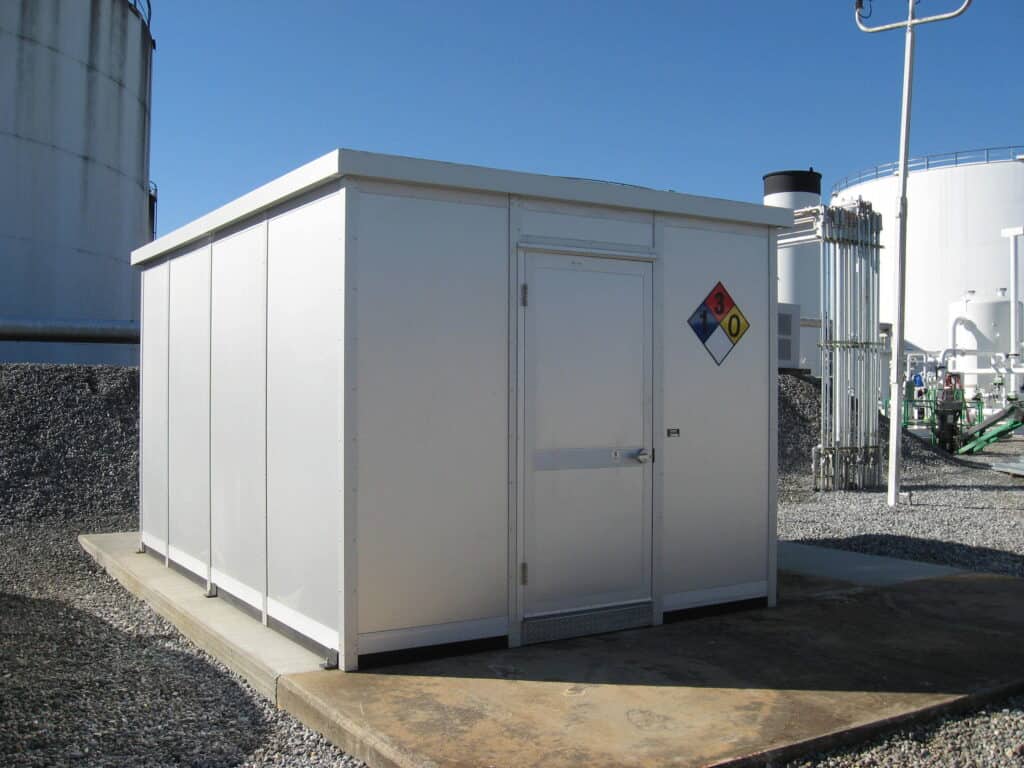 A small, white prefabricated industrial storage building with a single door, located on a gravel surface next to large storage tanks, with hazard signs on one side.