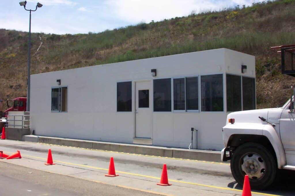 A white rectangular portable scale house with several windows, stationed by the roadside beside a hill. Orange traffic cones are lined in front, and a white truck is partially visible on the right.