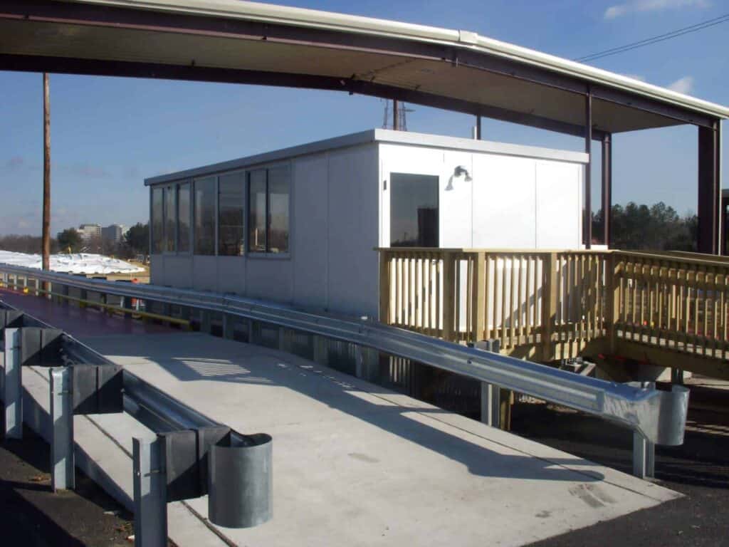 An accessible building with a ramp and railing at a public transportation station, featuring a covered walkway and benches on a sunny day with light snow on the ground, designed with innovative architecture.