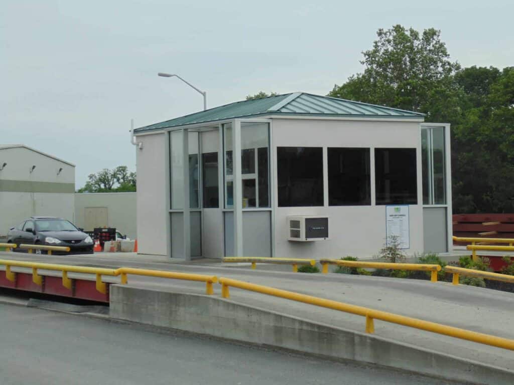 A security booth with large windows and a green roof at the entrance of a parking area, managed by property management, surrounded by yellow safety barriers. A car is parked nearby, with industrial buildings in the