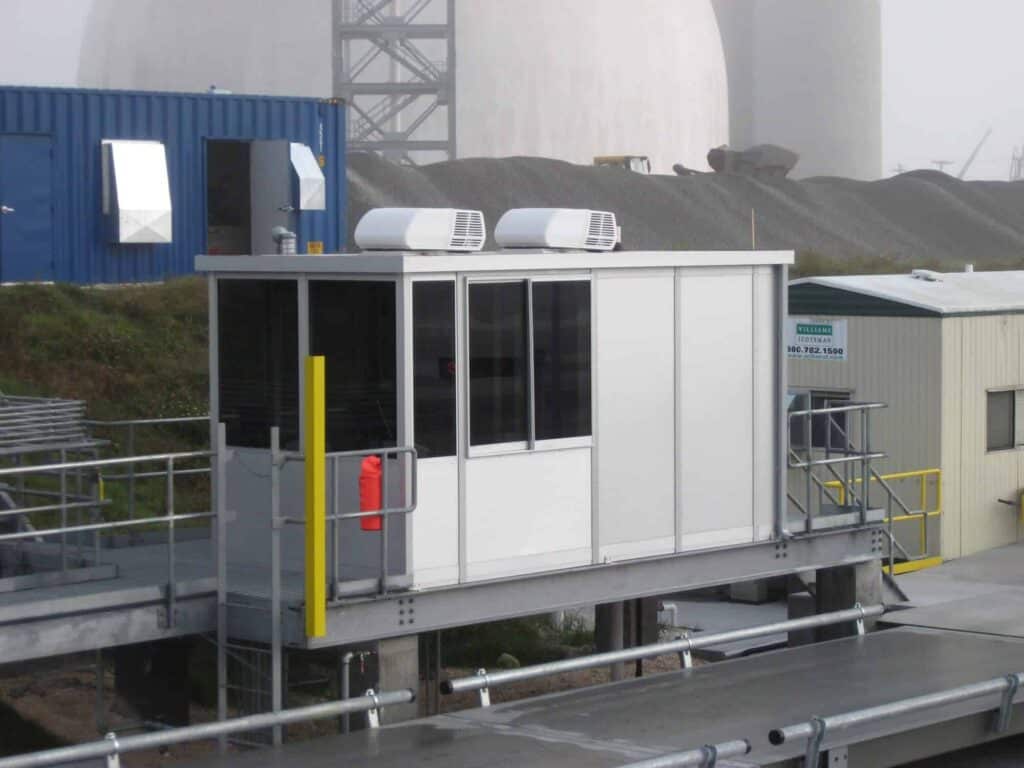 A small, modern portable office with air conditioning units on top, located on a metallic platform in an industrial setting with large structures in the background on a foggy day, often used for property management activities