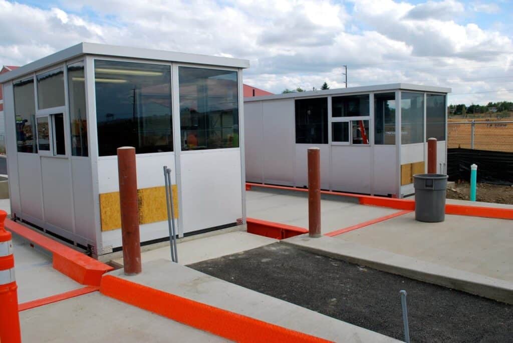 Two small, modern scale houses with glass windows and gray walls at a parking lot entrance, featuring bright orange barriers and clear skies.
