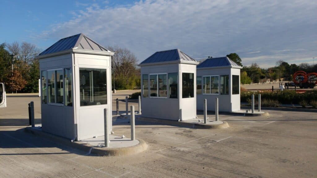 Two toll booths with gray and white paneling under a clear blue sky. The booths are surrounded by a large empty parking area with bollards in front of each booth.