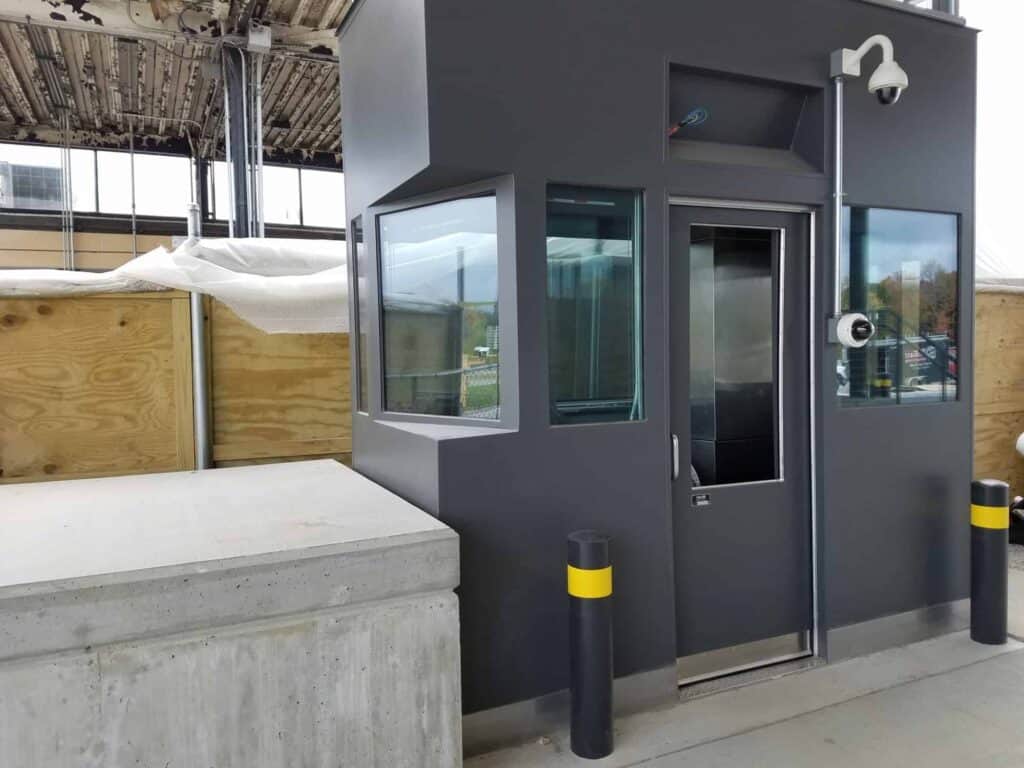 A modern toll booth security checkpoint building with a glass door, positioned next to a concrete platform and wooden barriers. Two security cameras are installed above the door.