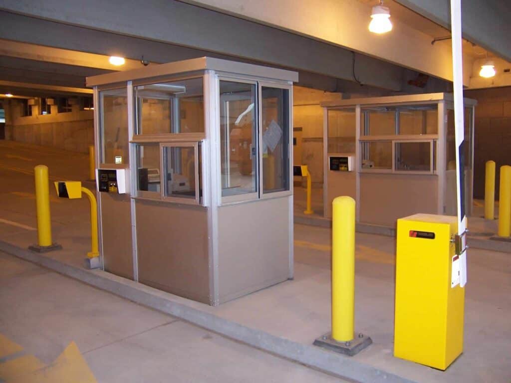 A security booth stationed at the entrance of a parking garage, flanked by yellow bollards and featuring a barrier gate to the right.