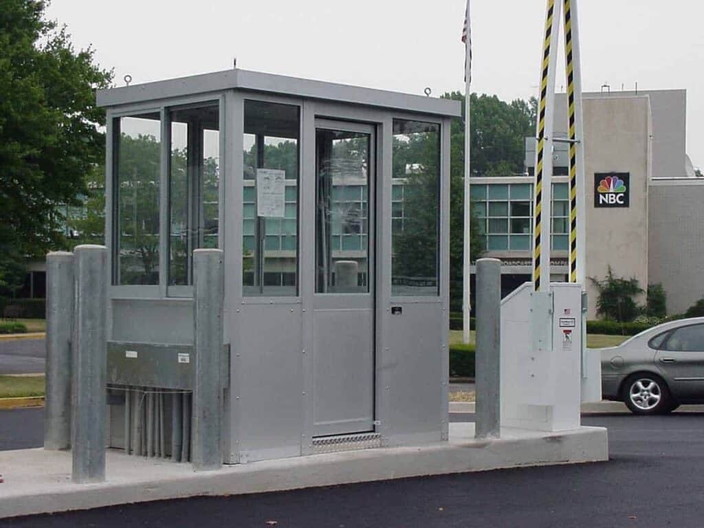 A security guard booth at the entrance of a parking facility with an NBC logo on a building in the background, a raised barrier arm, and a partially visible car.