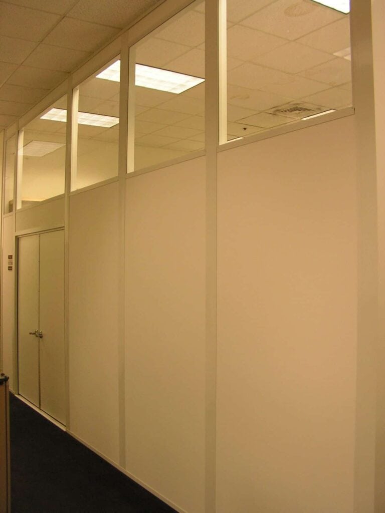 An office area with transparent glass walls above modular office partitions, a closed door, and carpeted floor, illuminated by soft lighting.