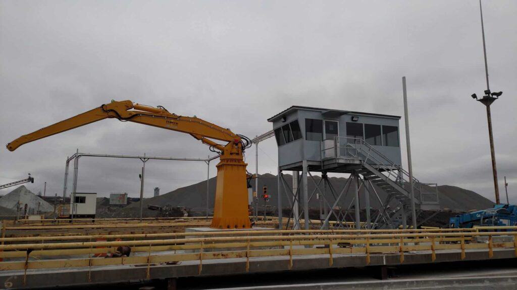 A large yellow industrial crane extending its arm over a pile of gravel, with operator booths in the background, under an overcast sky.