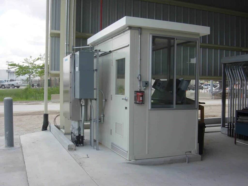 A security booth with large windows, positioned next to a building under an overhang. Behind the booth is a mechanical unit, and on the right is a gated entry system. This structure serves as one