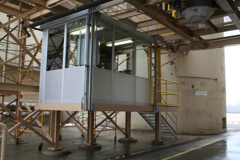 A small, elevated operator booth with glass windows, surrounded by metal railings, located inside an industrial building with visible support structures.