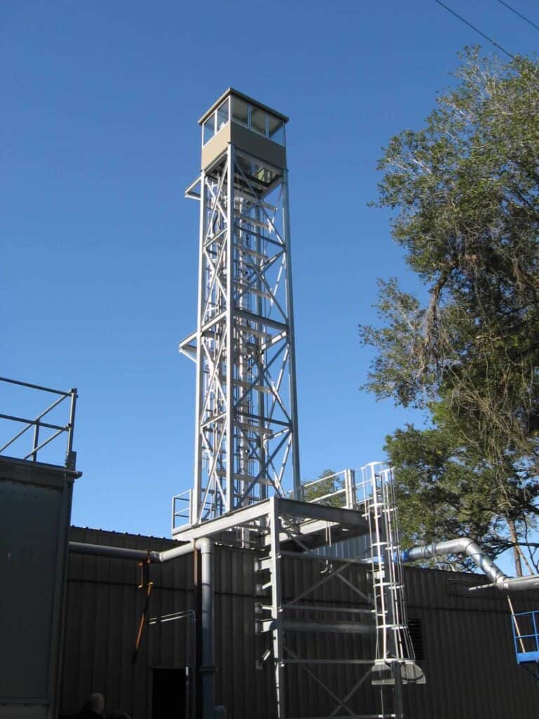 A tall metal guard tower with operator booths, surrounded by industrial structures and trees under a clear blue sky.
