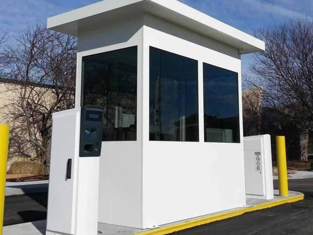 A small white parking booth with large glass windows, situated in a parking lot, equipped with air conditioning and security technology.