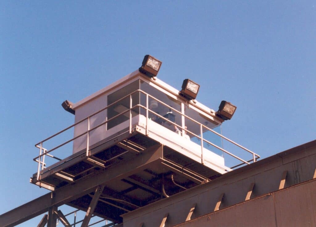 A small, elevated operator booth with a white facade and large windows sits atop a steel structure, under a clear blue sky. It is equipped with four large exterior lights.