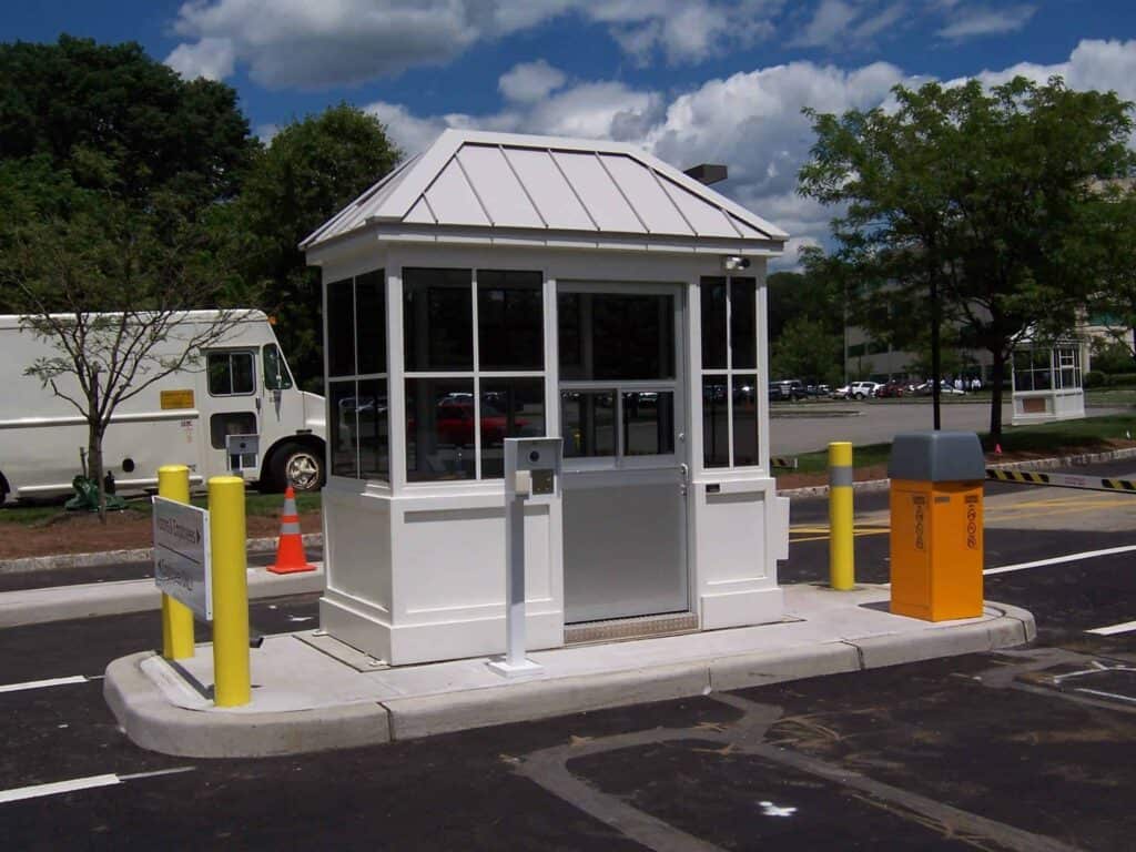 A white parking booth with a gabled roof, positioned at a parking lot entrance. There are yellow bollards around it, and a gray barrier gate. The background shows a truck and trees under
