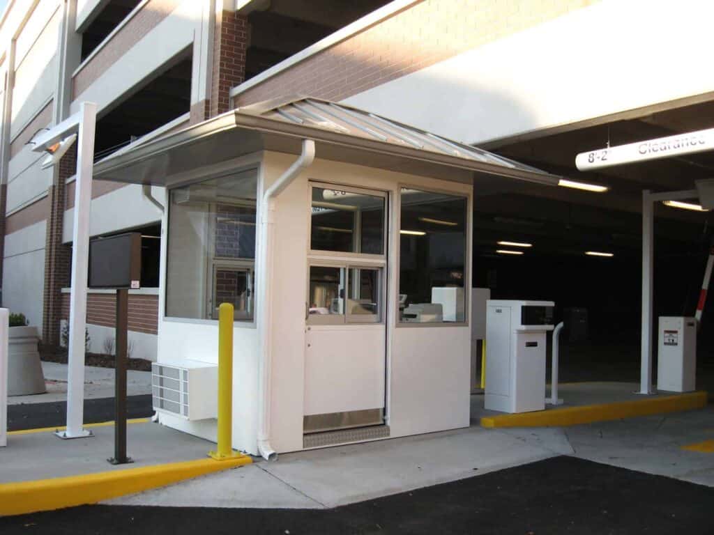 A parking lot entrance with a small parking booth equipped with monitors and communication devices, located under a covered driveway of a building complex.
