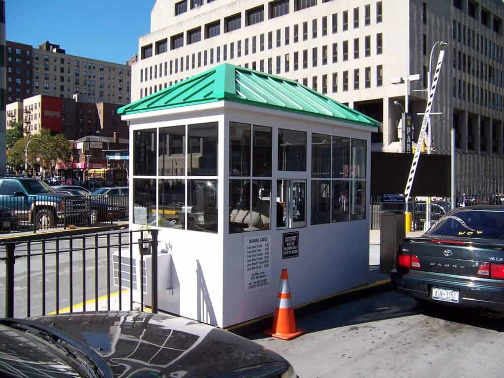 A small white parking booth with a green roof, surrounded by vehicles and traffic cones, located at an urban street setting with tall buildings in the background.