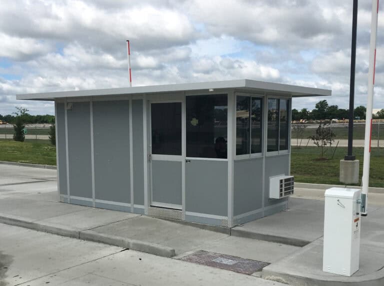 A small, modern security booth made of gray panels with large windows and equipped with a wall-mounted HVAC, located at the entrance of a parking area under a partly cloudy sky.
