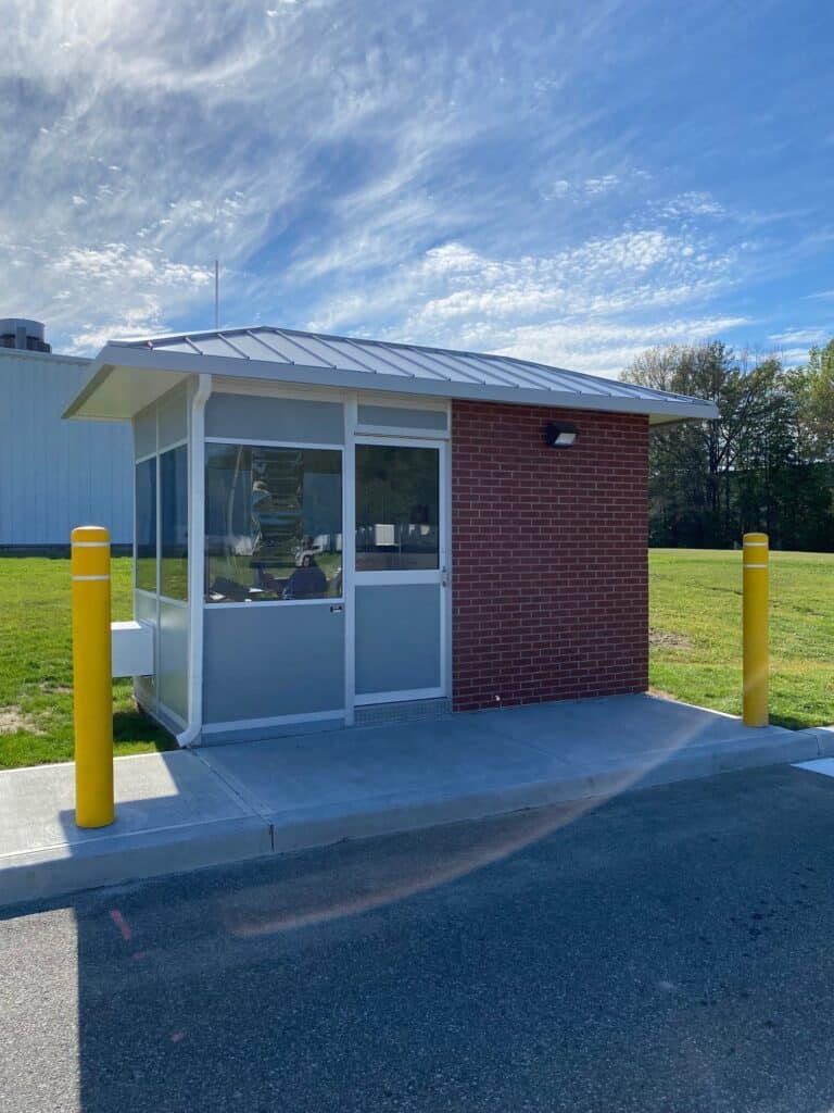 A small, modern security booth with factory-installed brick and glass walls, flanked by yellow bollards under a clear blue sky. There's grass and part of another building in the background.