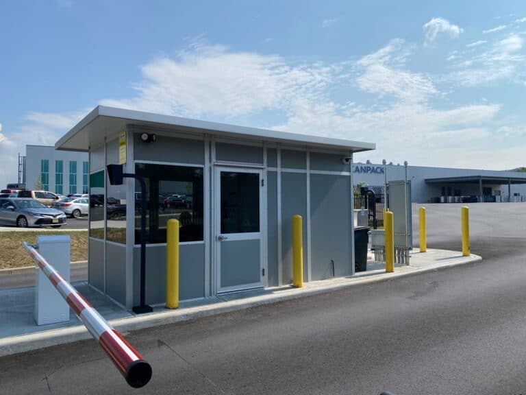 A modern security checkpoint with a lift gate at an industrial facility entrance features a roof overhang. The booth is gray with glass windows, surrounded by yellow bollards, under a clear blue sky.