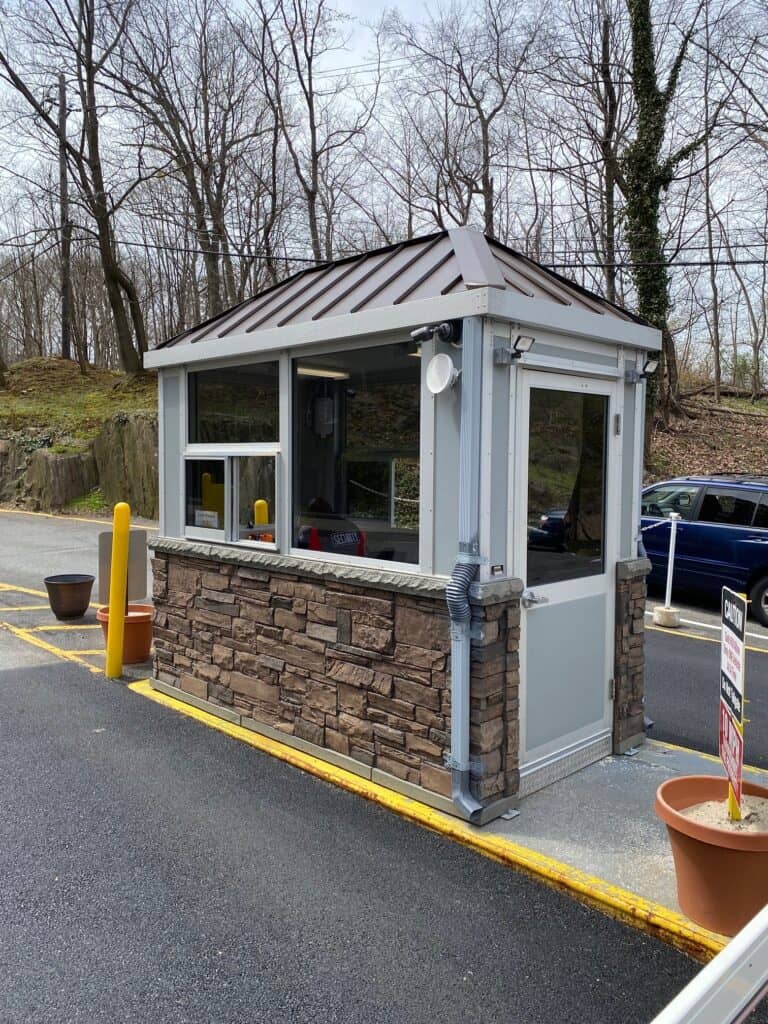 A small, sturdy parking attendant booth with faux stone and gray paneling, featuring wide windows and situated on an asphalt lot with a few cars and leafless trees in the background.