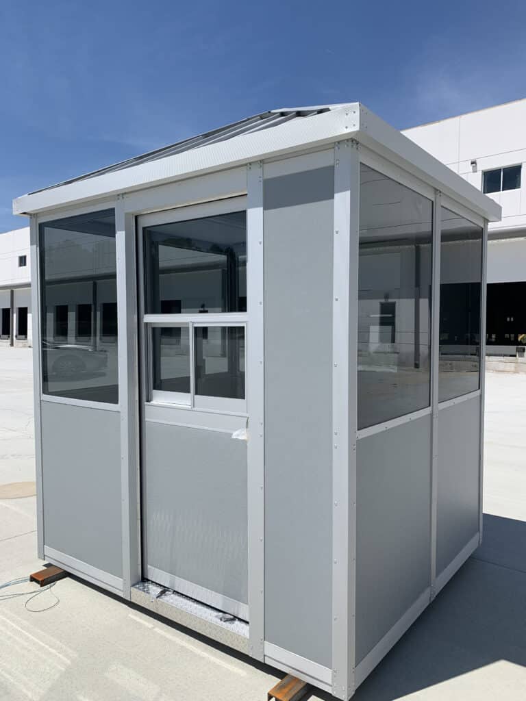 Small, standalone gray security booth with a heavy-duty aluminum transaction window installed on a concrete surface under a clear sky, reflecting the exterior building off its glass panels.