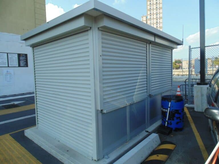 A small, rectangular parking attendant booth made of gray metal panels with closed security shutters, situated in a sunlit parking lot with nearby buildings visible in the background.