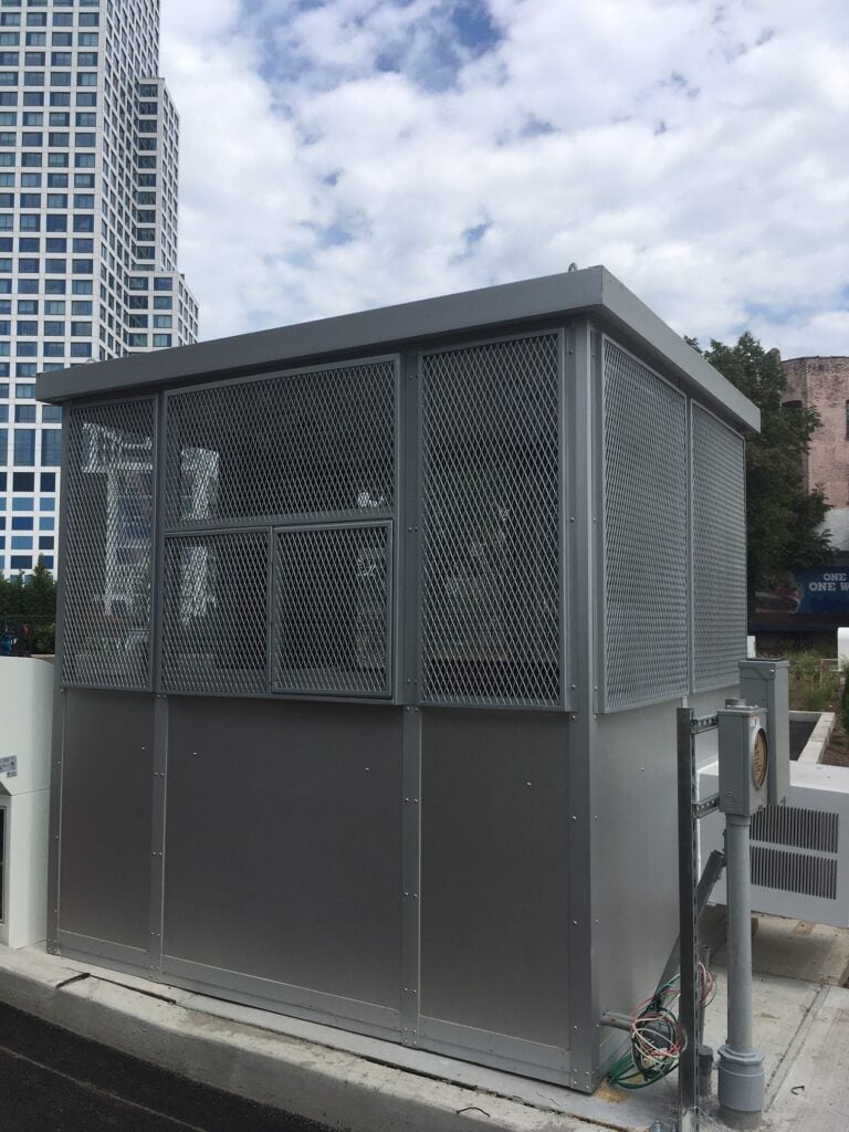 A metal utility enclosure with security screens, situated outdoors in an urban area with skyscrapers partly visible in the background under a cloudy sky.