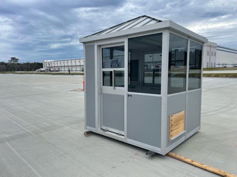 A small, gray security booth with large windows and a white metal roofing, situated on an expansive concrete lot under a cloudy sky.