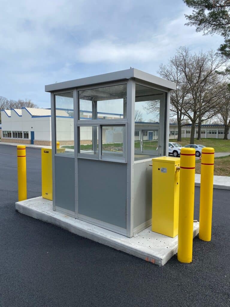 A small, modern gray security booth with a transaction window and glass windows, surrounded by yellow bollards on a parking lot, with buildings and trees in the background under a cloudy sky.