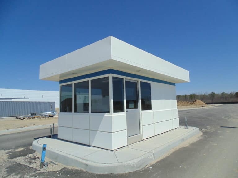 A small, modern guardhouse with a large aluminum sliding door and a flat roof, situated on a concrete base at the corner of a paved area, with a clear blue sky above and