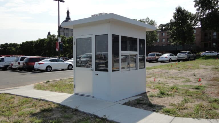 A small, modern security booth with large windows and a heavy duty aluminum swing door, installed on a concrete sidewalk, overlooking a parking lot filled with cars and a building in the background.