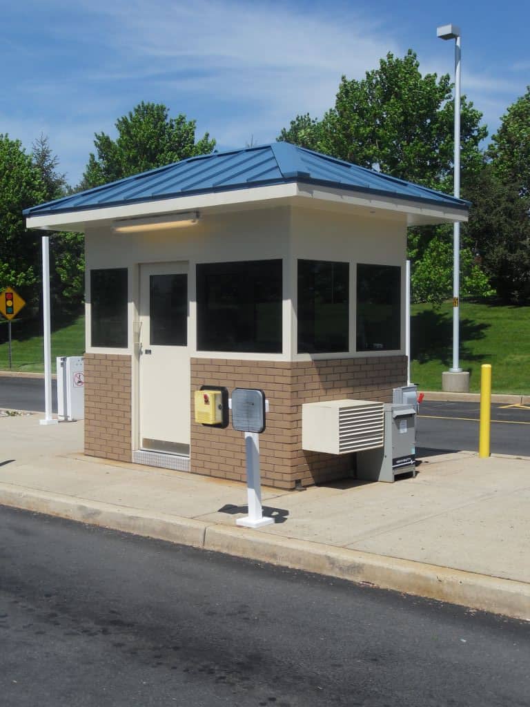 A small security booth with factory installed brick walls and a blue roof, situated in a parking lot. It features a large window, an air conditioning unit to the side, and is surrounded by lush green